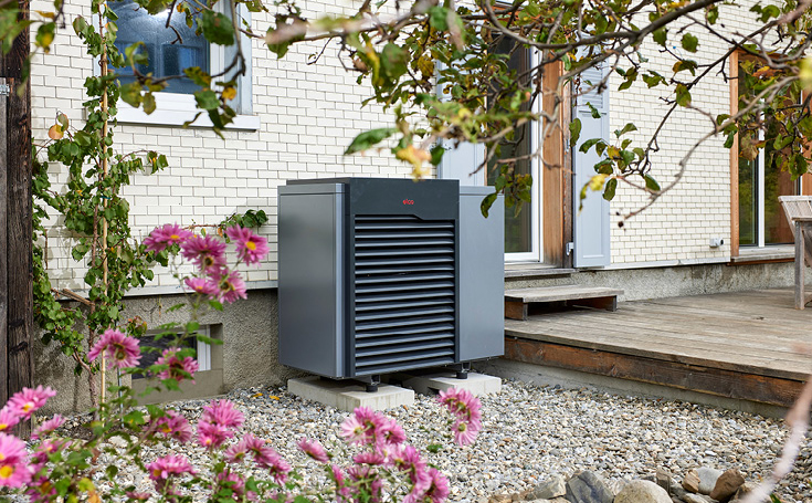 Outdoor-Wärmepumpeneinheit neben einem Haus mit weißen Ziegelwänden, umgeben von einem Garten mit rosa Blumen und grünem Laub.